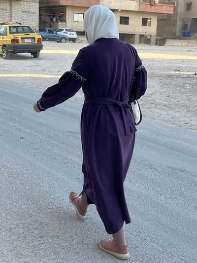 An individual is depicted walking away from the camera on a light gray paved road in Mu'adammiyat ash-Sham, Syria. The person wears a white hijab, a long-sleeved dark purple dress with a self-tie belt knotted at the back, and light pink sneakers. The dress features white zigzag stitching details on the sleeves. A white shoulder bag with a black strap is carried on the individual's right side. In the background to the left, a yellow sedan with a "TAXI" sign on its roof is parked, partially visible. Behind the vehicle, a light-colored, multi-story building with several windows is present. Further back and to the right, a blue pickup truck and a white SUV are visible on a dirt or gravel area. Multiple other buildings, appearing residential and varying in color and state of completion, line the distant background. The ground beyond the paved area consists of dry, dusty earth. The scene is illuminated by natural daylight.