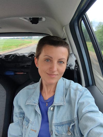 A woman with short brown hair and blue eyes is sitting in the passenger seat of a car, taking a selfie. She is wearing a light blue denim jacket over a royal blue t-shirt and a delicate gold necklace. Her left earlobe has a small, light blue stud earring. The interior of the car is visible, including the headrest and a dark bag in the back seat. Through the open car window on the right, a blurry, green landscape with trees is visible, indicating the car is in motion. The background outside the front windshield shows a road and a field with a sign in the distance that appears to read "Крывасельскі сельскі Савет" (Kryvaselski Village Council) in Belarusian Cyrillic. The sky is overcast.