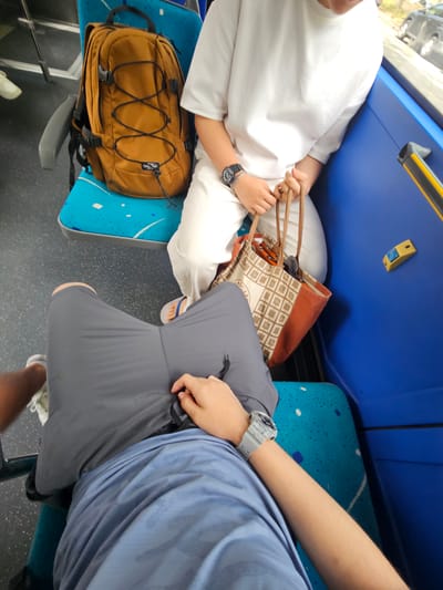 The image shows two people seated on a bus. The person in the foreground is wearing a blue t-shirt and grey shorts, with their legs crossed. The person behind them is wearing a white t-shirt and white pants, holding a patterned tote bag. A brown backpack is visible on a blue seat next to them. The bus interior features blue seating and a grey floor with white speckles. Outside the window, a car is partially visible, suggesting an urban environment. The scene appears to be a candid snapshot of everyday travel.