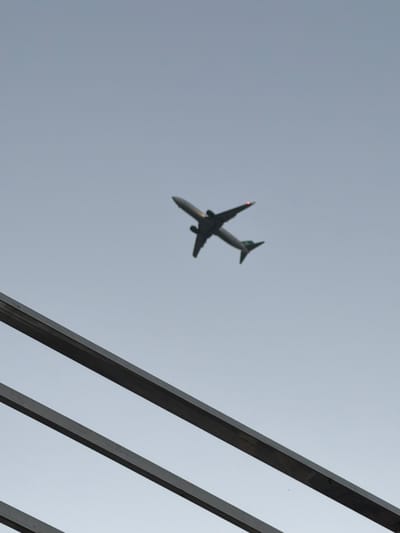 The image shows an airplane in flight against a light grey sky. The airplane is a passenger jet, silhouetted against the sky. It is angled downwards and to the left, with its wings and tail clearly visible. A small red light is visible on the tip of one of the airplane's wings. The sky is overcast, suggesting cloudy or hazy weather. In the foreground, there are several dark, angular structures that appear to be part of a building or a bridge. These structures are out of focus and run diagonally across the right side of the frame. The image was taken from a low angle, looking up at the sky and the airplane. There is no visible text in the image. The context provided suggests the location is Delhi, India, but there are no specific visual cues to confirm this. The time of day is likely daytime, given the brightness of the sky.