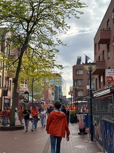 This image captures a lively street scene in a Dutch city, likely Eindhoven, during what appears to be a King's Day celebration, indicated by the widespread use of orange clothing among the crowds. The setting is a pedestrian-friendly street lined with brick buildings of varying architectural styles.
In the foreground on the left, a large deciduous tree with light green spring leaves partially frames the scene against an overcast sky with patches of brighter clouds. The street itself is paved with brick pavers.
Numerous people are present, most of whom are wearing vibrant orange jackets, shirts, or accessories. They are gathered along the street, standing, walking, and socializing, creating a festive atmosphere. On the right side of the street, temporary metal barriers are set up. Behind these barriers, a large, red-and-white striped barrel, labeled "JUMBO" (a Dutch supermarket chain), is filled with what appears to be dark fruit or berries. Further down the street, more people in orange are visible, suggesting a larger gathering or event area.
Several streetlights with large, opaque globes are positioned along the street. The buildings feature various commercial establishments. Visible text on signs includes: "美 安" and "MAY AN KAPSALON" (likely a hairdresser), "Ria Money Transfer," "Uber Eats," "PIOEN," and "ASPACT." In the background, modern glass buildings stand alongside older brick structures. The time of day appears to be daytime, with soft, diffused lighting. The overall impression is one of a vibrant public holiday celebration.