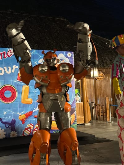 An indoor or covered outdoor scene depicts a person in a detailed orange and silver robot costume standing center. The robot has large, segmented arms raised upwards, with metallic gray claws. Its body is a combination of orange plating and gray mechanical components, featuring a rounded head with visible optical sensors. To the left of the robot, a blue backdrop displays a pattern of water and pool-themed graphics, including a large inflatable watermelon ring and the letter 'L'. Other smaller graphics on the backdrop include flowers, a water gun, and flip-flops. To the right of the robot, a person in a colorful, jester-like costume with a striped hat and diamond-patterned pants is visible. Behind the subjects, a wooden structure with a lit lantern and a sign that reads "SOÁT VẼ - Ticket control" is present. The ground is paved with gray tiles, and a dark, raised platform is visible in the foreground. The overall setting appears to be an entertainment or event venue, possibly a theme park or resort, in Hoa Lư Ward, Vietnam, suggested by the Vietnamese text on the sign.