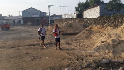 Two uniformed young men walk on a dirt road, past piles of sand and debris. Buildings line the background, some with corrugated metal roofs. A small truck is visible on the left, and power lines stretch across the sky. A stone wall runs along the right side of the image, separating the road from a metal-sided structure. The overall scene suggests a construction or improvement project in progress.