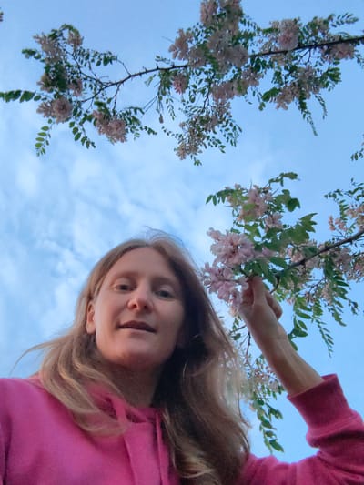 A young woman with blonde hair is captured from a low angle, looking upwards at the sky and a flowering tree. She is wearing a bright pink hooded sweatshirt. Her hand is reaching up to touch a cluster of pale pink flowers on a branch that extends over her. The sky is a soft blue with scattered white clouds. The scene is set outdoors, possibly in a park or a natural area within the Зуйское сельское поселение, Ukraine. The overall mood is serene and suggestive of a pleasant spring day.