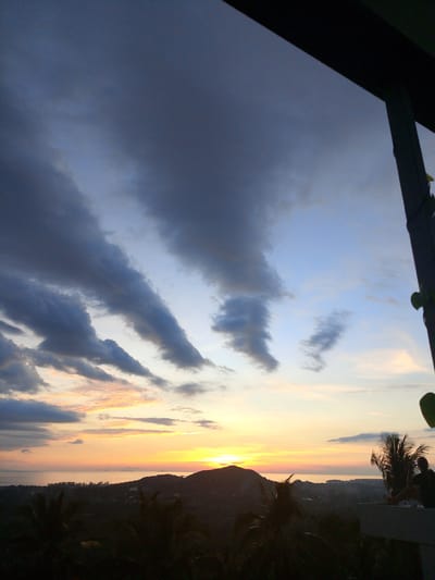 A wide-angle, low-light, high-angle shot captures a dramatic sunset over the ocean and hilly landscape of Ban Tai Subdistrict, Thailand. The sky is dominated by a gradient of blues and grays, with streaks of dark, wispy clouds stretching across the frame. The lower portion of the sky transitions into a vibrant band of orange and yellow hues where the sun is setting, illuminating the ocean and the silhouetted outlines of distant islands and the coastline.

In the foreground, a dark, silhouetted structure, possibly a balcony or roof overhang, occupies the top right corner, partially obscuring the sky. Below this, dark silhouettes of palm trees and vegetation create a textured foreground. To the right, a dark silhouette of a person is seated on the edge of what appears to be a concrete structure, facing towards the ocean and the sunset. The person is seated with their legs bent and their arms resting on their knees, appearing to be observing the view. The overall scene suggests a peaceful, contemplative moment during twilight.