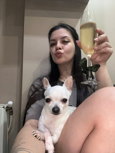 A woman with dark hair is holding a champagne flute filled with a pale yellow liquid, a dark green ribbon tied around the stem. She is puckering her lips as if for a kiss. A small white Chihuahua is resting on her arm. The woman's left arm is bare, showing a tattoo on her forearm. She is wearing a gray t-shirt. The background appears to be an indoor setting with neutral-colored walls. A radiator with pipes is visible at the bottom of the frame. The image is taken in portrait orientation. The lighting suggests it could be daytime or a well-lit indoor space. The overall mood is casual and celebratory.