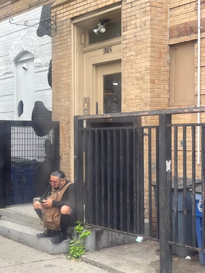 The image depicts a street scene in New York City, likely during the daytime, given the lighting. A person is squatting on a set of concrete steps next to a building. The building's facade is made of light-colored brick, with a door labeled "1206" visible. There's a black metal gate in front of a section of the building.
The person is wearing dark clothing, including ripped pants, a black long-sleeved shirt, and black boots. They have a tan messenger bag slung over their shoulder and are wearing earbuds. Their facial expression is neutral or perhaps weary. Small green plants are growing from a crack in the concrete near the steps.
In the background, through the metal gate, a blue recycling bin and a dark gray trash can are visible. The ground is paved with concrete. The overall atmosphere appears to be urban and somewhat gritty, a common characteristic of street scenes in many parts of New York. No specific event is clearly taking place, other than the individual resting on the steps.