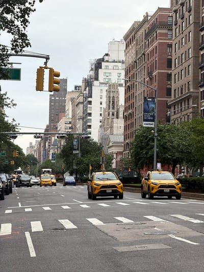 This is a medium-angle, eye-level, landscape photograph taken from the perspective of someone standing on a sidewalk or crosswalk, looking down a city street. The image is oriented vertically, with the top of the buildings appearing on the left side of the frame. The scene depicts a bustling street in New York City, with several yellow taxis driving down the road. The buildings on either side of the street are tall and made of brick and stone, characteristic of early 20th-century architecture. Trees line the sidewalks, adding greenery to the urban environment. A traffic light is visible in the foreground, and a pedestrian crossing is marked on the asphalt. The sky is overcast, suggesting a cloudy or hazy day. Banners are hung on poles along the street, with one clearly displaying the text "NYC FINANCE STATE OF MIND CANTOR". Other visible text includes "ONE WAY" and license plate numbers like "2J77", "5C82", "Y2067540", and "Y2067510". The overall impression is of a typical day in a major metropolitan area, with urban life in full swing.
