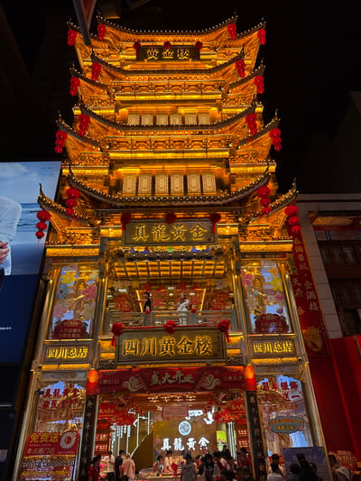 Here's a forensic description of the image:
**Scene Overview:** The image depicts an elaborate, multi-tiered, and brightly lit structure, predominantly golden in color, adorned with red lanterns and intricate decorations. The structure is heavily ornamented with signage in Chinese characters. To the right of the golden structure is an area with people.
**Location:** The presence of Sichuan business branding suggests that this location is likely a retail or commercial district in Jinjiang District, China.
**People:** In the background, there is a glimpse of a crowd of people.
**Subjects:** The main subject is the illuminated golden structure. The structure's architecture and signage imply a business establishment or a decorative display.
**Objects:** Prominent objects include the golden structure, numerous red lanterns, signage with Chinese characters, a television screen at the base of the structure displaying a person, and potentially, other commercial elements in the surrounding area.
**Actions:** The primary action is the visual display of the elaborate golden structure, presumably designed to attract attention and convey a sense of prosperity or celebration.
**Interactions:** The interaction is primarily between the structure and the environment, with the lighting highlighting its details and attracting attention from passersby.
**Environment:** The environment appears to be an urban setting at night. The presence of lights and potential commercial elements suggests a busy area within Jinjiang District, China.