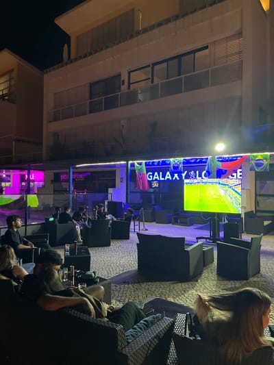 The image shows an outdoor seating area at night, likely a lounge or cafe, with several people seated and watching a large screen displaying a soccer match. The setting appears to be in Lisbon, Portugal, judging by the flags displayed, which include the Portuguese flag and the flag of Brazil. The lounge's name, "GALAXY LOUNGE," is visible on a lit sign above the screen. The seating area is furnished with dark rattan chairs and tables, and the ground is paved with cobblestones. Some people are smoking shisha, indicated by pipes on the tables. The overall atmosphere is casual and relaxed, with the focus of attention being the soccer game.