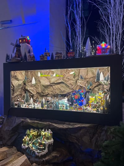 This image depicts a multi-level miniature Christmas village display located in Sélestat, France, presented in a dark interior environment with ambient lighting.

The central feature is a large, rectangular display box, framed in black, containing a miniature winter village scene. The interior background of this box is composed of crumpled brown paper, simulating rocky terrain or mountains, with sparse white elements representing snow. Within this scene, numerous miniature buildings, including houses, shops, and a church, are covered in artificial snow and illuminated with integrated lights. Miniature evergreen trees and bare white trees are interspersed throughout the village. A prominent, illuminated blue Ferris wheel with red accents is visible on the right side of the main village. Tiny figures are positioned to suggest activities such as walking and skating. A black power cord is visible extending from the lower left of the display box.

Above this main display, a second tier of miniature elements is present. On the left, a miniature windmill and a house perched on a snowy hill are visible. A ski lift system with miniature red and yellow/white cabins traverses horizontally across this upper tier. On the right side of the upper tier, additional miniature houses and a brightly lit red barn-like structure are situated among dense white, bare tree branches, some illuminated with small lights, simulating a winter forest. A strong blue light source illuminates the wall behind the upper left portion of this display.

Below the main display box, the base is covered with dark, crumpled material, mimicking rugged terrain. Logs are visible on the far left. A smaller, illuminated miniature scene depicting figures ice skating on a pond, surrounded by snow-covered buildings and trees, is positioned centrally on this lower level. A dark green artificial pine branch is visible on the far right, near the base of the display. The background beyond the display appears dark, possibly a window reflecting some lights from the scene.