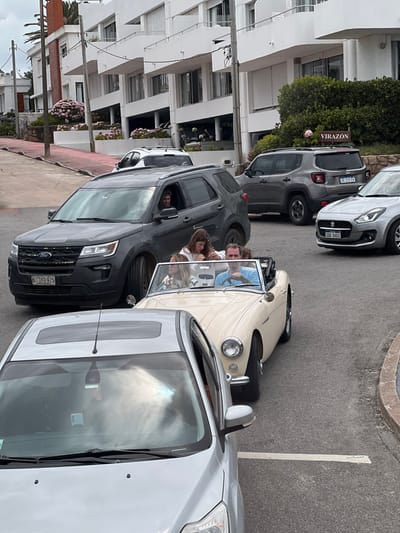 The image depicts a bustling street scene in what appears to be a coastal or resort town, consistent with Punta del Este, Uruguay. Several cars are visible, either parked or temporarily stopped on an asphalt road alongside modern multi-story buildings.
In the foreground, a prominent light-colored, possibly cream or beige, vintage convertible car, resembling a classic roadster, holds four cheerful individuals. A man is seated in the driver's side, looking ahead, while three women are comfortably seated in the back, all appearing to be enjoying the moment with relaxed expressions.
Adjacent to the convertible on the left is a dark grey Ford SUV, with a person visible in the driver's seat. Its license plate reads "G 399475." Further back, a dark grey SUV similar to a Jeep Renegade is parked, displaying the license plate "AB 237 EX." A light grey hatchback, with license plate "SBA 5653," is also visible in the mid-ground. Part of a silver car is seen in the immediate right foreground.
The background is dominated by contemporary white buildings featuring multiple stories, balconies, and large windows, some fitted with rolling shutters. Green foliage, including bushes with vibrant pink flowers, decorates the area in front of the buildings and along a retaining wall. A sign bearing the text "VIRAZON" is clearly visible on one of the buildings, likely indicating a hotel or residential complex.
The scene unfolds during bright daylight hours, suggesting pleasant weather conducive to an open-top car ride. The overall impression is one of leisure and activity in an urban or holiday setting.