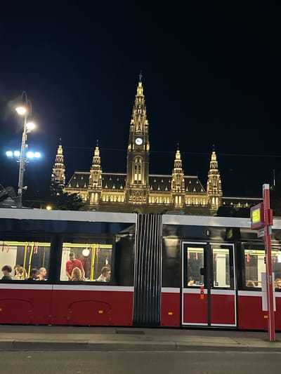 The image captures a night scene in Vienna, Austria, focusing on the Vienna City Hall (Rathaus) and a red and black tram passing in front of it. The City Hall is a large, ornate Gothic Revival building, brightly illuminated and detailed with spires, arched windows, and a prominent clock tower. The tram, with its distinctive red lower half and black upper section, has its windows revealing several passengers inside. The tram is stopped or moving slowly, positioned on a paved street. Streetlights are visible, casting a warm glow. The sky is dark, indicating nighttime. The overall impression is of a well-lit urban environment at night, with the historic architecture of the City Hall serving as a dramatic backdrop to the modern public transportation.
