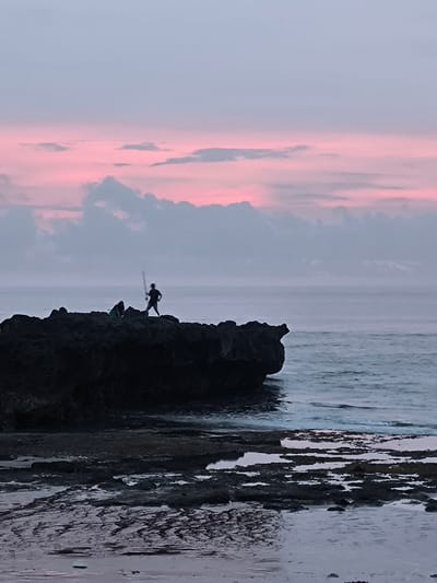 The image depicts a coastal scene in Canggu, Indonesia, at dusk or dawn, evidenced by the pink and grey sky. A dark, rocky outcrop is positioned in the middle ground, extending into the sea. Two figures are silhouetted against the sky, standing on the rocks. One is holding a long object, possibly a fishing rod or spear. The foreground features a wet, rocky beach area with shallow pools reflecting the sky. The sea is visible in the background, with gentle waves breaking.