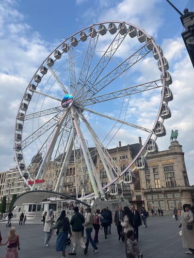 The image shows a large Ferris wheel with gray and white cabins against a blue sky with white clouds. The Ferris wheel is partially obscuring a building with classical architectural details, including arched windows and decorative elements. A statue of a figure on horseback sits atop the building. People are walking on a paved surface in the foreground, appearing as a blurred row. The location is Antwerp, Belgium, as "Antwerp's Wheel" is written on the center of the wheel.