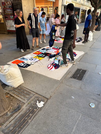 In Seville, Spain, on a sun-drenched paved street, a street vendor, a Black man wearing a black t-shirt and gray cargo pants, displays a collection of soccer jerseys. He holds up a blue and yellow jersey with the number 10 and a Brazilian flag. Laid out on a white cloth on the ground are numerous other jerseys in various colors and designs, some neatly folded, others in a pile. A large white plastic bag, possibly containing more merchandise, sits to the left of the display. To the right, another man, dressed in a blue t-shirt and black shorts, stands near a tree. Several other people, including women in dresses and a boy in a blue and white striped shirt, are seen browsing or walking in the background. The street appears to be a commercial area, with shop facades visible behind the people. The ground is paved with granite slabs and includes a metal grate for drainage.