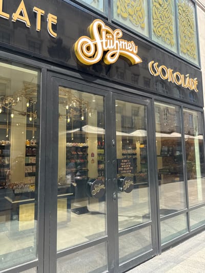 The image shows the exterior of the Stühmer chocolate shop on Andrássy Avenue in Budapest, Hungary. The shop has a large glass storefront with double doors. Above the entrance, the Stühmer logo is visible, as well as the word "CSOKOLÁDÉ" (Hungarian for chocolate). Inside the shop, display cases filled with chocolates and other confectionery are visible. The street outside is paved with cobblestones, and the buildings across the street are also visible. The lighting suggests it is daytime.