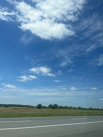 The image depicts a wide, open landscape under a vast blue sky with scattered white clouds. In the foreground, a paved road with white lane markings stretches horizontally across the frame, suggesting a highway or a similar thoroughfare. Beyond the road, a field of dry, golden grass extends towards a line of trees that forms the horizon. The trees are varied in height and density, creating a natural border between the field and the sky. The overall impression is one of a rural or semi-rural setting in Balivka, Ukraine, characterized by open spaces and natural elements under a bright, partly cloudy sky. There are no people or discernible actions visible in the scene.