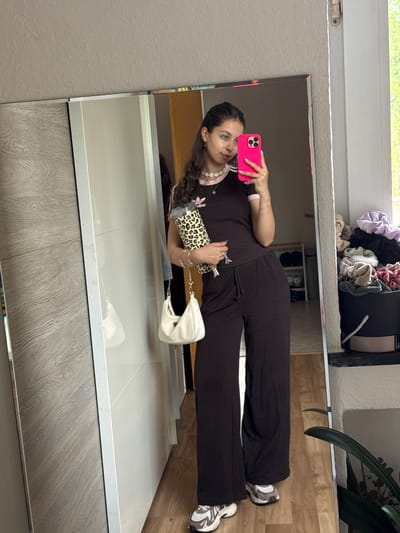 A young woman is taking a selfie in a mirror. She is wearing a brown Adidas tracksuit, a shell necklace, and white sneakers. She is holding a pink iPhone in her right hand and a leopard-print thermos and a white handbag in her left hand. The mirror is reflecting a white tiled shelf and a wooden floor. In the background, there is a bucket filled with hair scrunchies and a green plant. The photograph was taken in an unknown location in Bruchköbel, Germany.