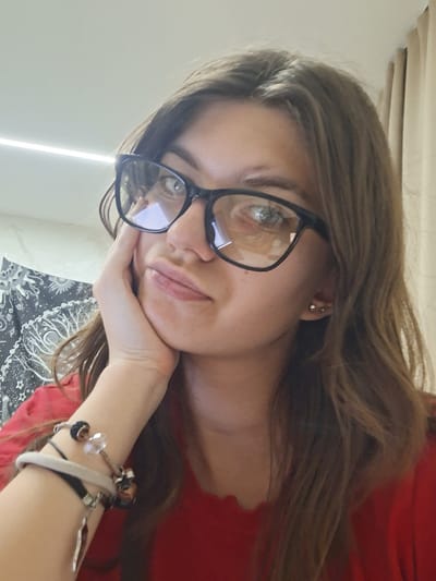 A woman with long brown hair and glasses is looking at the camera. She is wearing a red shirt and has several bracelets on her wrist. Her head is resting on her hand. A black and white patterned blanket is visible in the lower part of the frame. A light beam is visible on the left side of the image. The location is unknown, and there is no evidence to suggest it is Panagyurishte, Bulgaria.