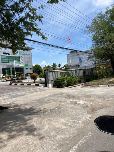 The image shows the BPJS Ketenagakerjaan office in Langsa, Indonesia. The building is modern with large windows and signage. In front of the building, there is a paved area with some landscaping, including bushes and small trees. A gray fence with decorative elements runs along the property line. The Indonesian flag is flying from a flagpole on the right side of the image. Overhead, numerous electrical wires are visible against a blue sky with scattered clouds. The overall scene suggests a functional government office in an urban or suburban setting.