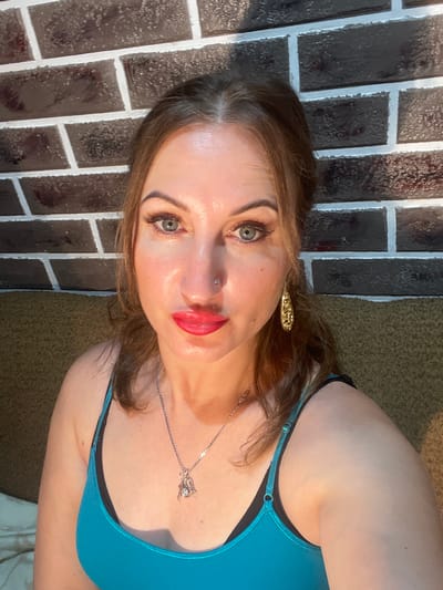 This is a medium shot, selfie-style photograph taken indoors. The subject is a woman with brown hair, green eyes, and full lips painted with red lipstick. She is wearing a teal camisole with black bra straps visible underneath, and a silver necklace with a pendant. A large, ornate gold earring is visible on her right ear. She has a nose ring. The background consists of a brick wall and a textured couch. The lighting suggests a warm, possibly artificial, light source illuminating the subject from the side. The image does not provide any specific details to locate it in Zarichne, Ukraine.