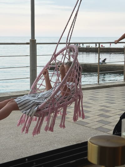 In Partenit, Ukraine, a young girl is sitting in a pink macrame hammock chair, suspended by ropes, with a view of the sea. She is wearing a striped dress and appears to be looking down or to the side. The hammock is adorned with pink fringe. The background features a pier with several people fishing, and the tranquil sea under a cloudy sky. The foreground shows a tiled walkway and a portion of a table with a metallic object, possibly a candle holder or a part of a lamp.