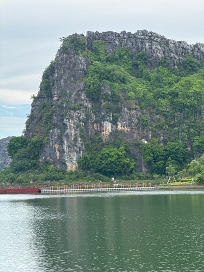 An overcast day frames a steep, rocky karst mountain covered in lush green vegetation. The mountain dominates the upper half of the image. In the foreground, a calm, green body of water reflects the sky and the verdant hillside. Along the edge of the water, a man-made walkway extends across the scene. This walkway is supported by a series of white floats and is lined with orange railings. A red barge is docked to the left of the walkway. Two people are visible on the walkway, appearing to be walking. The location is Hoa Lư Ward, Vietnam, a region known for its dramatic karst landscapes and waterways.