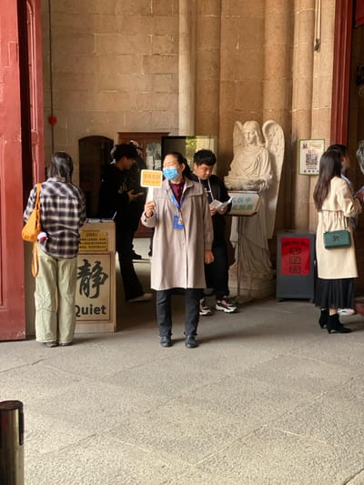 This image, taken in Guangzhou, China, depicts an indoor or covered outdoor area of a building, likely a temple, church, or historic site. Several people are present, appearing to be queuing or gathered respectfully.

In the foreground, a person with long dark hair, wearing a plaid shirt and light pants, stands with their back to the camera, carrying a light brown bag. Adjacent to them, a person in a black jacket is visible. The central figure is an attendant, dressed in a light trench coat, dark pants, and a blue face mask, holding a yellow sign that reads "保持安静" (Keep quiet) and "Keep quiet." Behind this attendant, another individual in dark clothing is partially visible. Further back, a young woman with long dark hair, wearing a light beige coat and black pleated skirt, stands near a grey "Offering Box" (奉献箱).

The setting features sturdy, light brown stone walls and a paved floor. A white angelic statue with wings is prominent behind the attendant. Next to the statue, a white sign states "礼仪中禁止拍照" (No photography during ceremony). On the ground, near the masked attendant, a wooden sign prominently displays the Chinese character "静" (Jìng - quiet/calm) and the English word "Quiet." The presence of an offering box and signs requesting silence and prohibiting photography during ceremonies suggests a place of worship or a site requiring solemnity. The activity appears to be an orderly visit or attendance at a quiet event. The scene is illuminated by natural, albeit somewhat dim, daylight.