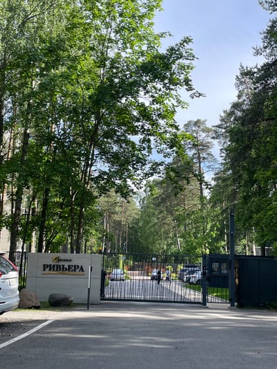 The image shows a gated entrance to a residential complex or facility called "Морская РИВЬЕРА" (Morskaya Riviera). The sign is large, light-colored concrete with gold lettering. Two dark grey boulders are placed in front of the sign. A white Volvo SUV is parked to the left of the sign, and a black metal gate is open, allowing passage into the complex. Through the gate, several cars and two people are visible on a paved driveway surrounded by green grass. The complex is situated within a wooded area with tall trees, suggesting it's located in Zelenogorsk, Russia, a resort town known for its pine forests and coastal environment. The sky is partly cloudy and the overall scene depicts a sunny day.