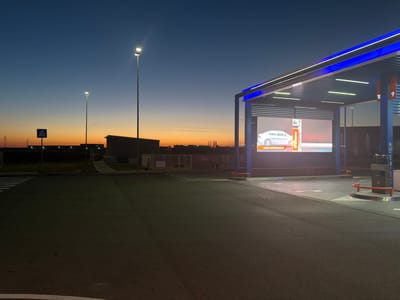 The image depicts an outdoor scene at twilight, likely in an industrial or commercial area within Stara Pazova Municipality, Serbia. A modern gas station or fueling station is prominently featured on the right side, illuminated with blue LED strips outlining the canopy. The station displays a large digital screen showing a car advertisement. Two tall lampposts illuminate the surrounding asphalt parking area. In the background, there is a smaller building and pedestrian crossing with a traffic sign. The horizon shows a faint orange glow, suggesting either sunrise or sunset. No people are visible in the image. The overall environment appears clean and well-maintained.