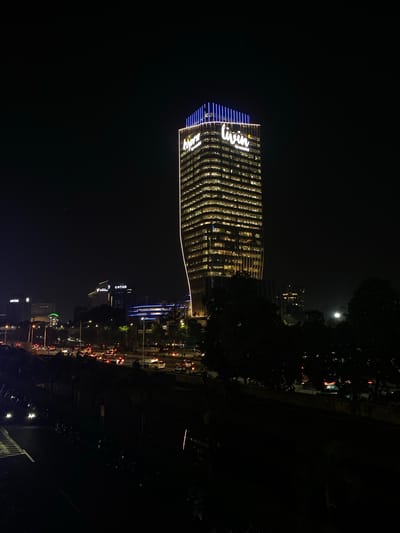 This is a nighttime, slightly rotated, vertical image of a modern, illuminated skyscraper in West Jakarta, Indonesia. The building, with its distinctive wavy facade, is lit from within, showcasing rows of brightly lit windows that create a dynamic pattern. The word "livin" is prominently displayed in white neon lettering across the upper part of the building, accented by blue lights along the edge.
The scene depicts an urban environment at night, with a full moon visible in the dark sky above the trees on the right side of the frame. In the background, other buildings and city lights are visible, suggesting a bustling cityscape. A busy street with car headlights and taillights is also apparent, indicating ongoing traffic. The overall mood is one of urban vibrancy and activity at night. The image is characterized by strong contrasts between the brightly lit building and the dark night sky and surrounding foliage.