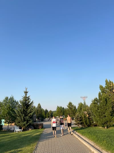 An outdoor scene captured in the city of Емельяновское озеро, Russia, depicts three young adult males walking away from the camera on a paved pathway under a clear blue sky. The pathway, constructed of grey rectangular pavers, extends into the distance, slightly curving left. A concrete curb separates the path from a manicured grass verge on the right.
From left to right, the individuals are dressed as follows: The first man wears a white sleeveless tank top with red and black lettering on the back (partially obscured) and black shorts, with light-colored footwear. The second man wears a black tank top with "JORDAN 23" printed in white on the back and light-colored patterned shorts, wearing white sandals. The third man is shirtless, wearing black shorts and dark sandals. All three men are walking in the same direction, with long shadows cast behind them by the sun, indicating a low sun angle (morning or late afternoon).
The environment is rich with vegetation. On both sides of the path, mature coniferous trees, appearing to be fir or spruce, are prominent, interspersed with lighter green deciduous trees. On the left side of the path, a white plastic lounge chair rests on the grass. In the middle distance, behind the trees on the right, a distinctive red and white metal structure, possibly a recreational or utility tower, rises above the tree line. The sky above is uniformly clear and blue, devoid of clouds. Additional people are faintly visible further down the path in the background.