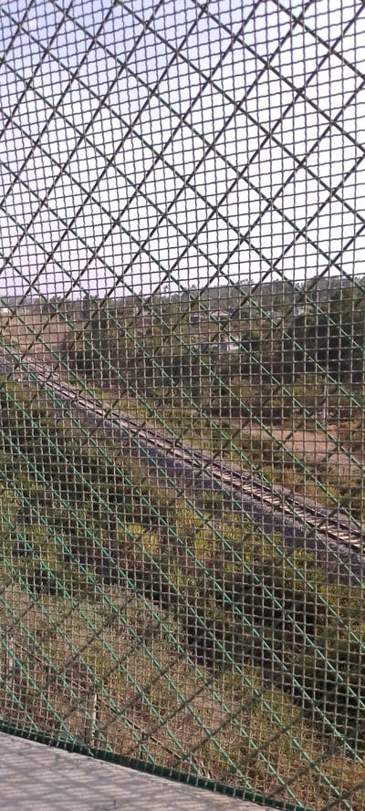 The image presents an outdoor scene viewed through a prominent wire mesh fence, which occupies almost the entire foreground. The fence, appearing to be made of green plastic-coated metal, forms a diamond-shaped grid pattern that covers the entire frame and is in sharp focus. The individual strands of the mesh are visible, creating a repetitive visual texture.

Beyond the fence, the landscape is visible but largely out of focus, suggesting a narrow depth of field with the fence as the primary subject. The perspective is from an elevated position, looking down onto a natural environment. In the mid-ground, a railway track can be seen curving gently from the lower-left portion of the image towards the center-right. The track consists of dark rails laid on light-colored sleepers and ballast, winding through the terrain.

The surrounding landscape is characterized by rolling hills or undulating ground covered densely with green vegetation, including trees, bushes, and grass. In the further background, nestled among the trees, a few light-colored buildings or houses are faintly discernible. The sky above is a light, possibly hazy or overcast, providing soft illumination to the scene. The overall lighting suggests it is daytime. No specific human activity or identifiable text is visible in the image.