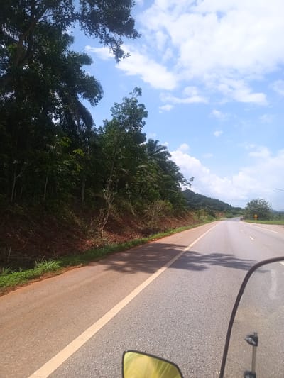 This image, likely taken in Plai Phraya, Thailand, depicts a paved road stretching into the distance. The road is flanked by lush greenery on one side and a clear sky with scattered clouds above. The viewpoint is from a vehicle, perhaps a motorcycle, as evidenced by the visible windshield and part of the mirror. The road markings are visible, indicating lane divisions. The vegetation appears dense and tropical. The image suggests a rural or semi-rural setting.
