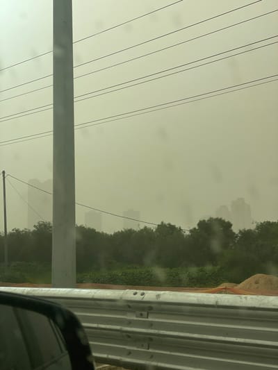 The image depicts an outdoor urban landscape in Ramat HaSharon, Israel, obscured by a dense, yellowish-brown atmospheric haze, characteristic of a dust storm, significantly reducing visibility. The photograph appears to be taken from inside a vehicle, indicated by a partial view of a dark car's side mirror and frame in the lower left, and faint circular smudges on what appears to be a window.
In the foreground, a metallic, multi-section guardrail runs horizontally across the lower portion of the frame, bolted together at regular intervals. Behind the guardrail, light brown earth and an orange construction safety net are visible, along with a small pile of sand to the right.
The midground features a line of dense, dark green trees and shrubs. A tall, light grey utility pole stands vertically in the left-center, extending towards the top of the frame. Multiple dark, thin utility lines stretch horizontally across the hazy sky, seemingly connected to this pole and other, less distinct structures. A smaller, darker utility pole is partially visible further left, carrying at least one wire.
In the background, the silhouettes of several multi-story buildings, consistent with an urban skyline, are faintly discernible through the thick haze. Their forms are indistinct and heavily blurred due to the atmospheric conditions. No individuals or explicit actions are visible within the frame.