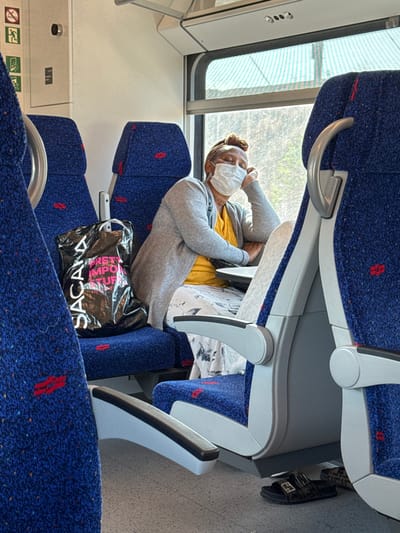 The image displays the interior of a modern passenger train carriage during daylight hours, consistent with Israel Railways' design, within the context of the Hevel Yavne Regional Council, Israel.
A light-skinned individual is seated in a window seat on the right side of the frame, appearing to be asleep or resting. They wear a white medical face mask, a mustard yellow top, a light grey open cardigan, and a white patterned skirt. Their hair is covered by a light brown headwrap. Their head is tilted to the left, supported by their right hand which rests on a small, light grey foldable tray table. On the floor beneath them, two sandals are visible: a black double-buckle slide and a patterned slipper.
To the left of the person, on the adjacent seat, a black plastic shopping bag with white text "SAYANA" and "PRETTY IMPORTANT STUFF" is placed. The train seats are upholstered in blue fabric with small red speckled patterns, framed by light grey and white plastic armrests and seat backs. Overhead luggage racks are partially visible. Through the window, bright sunlight illuminates a green and white striped structure or awning, suggesting the train is either stationary at a platform or passing industrial infrastructure. On the interior wall to the upper left, emergency exit signage and a circular call button are mounted. The train floor is light grey.