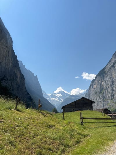 The image depicts a rustic wooden barn situated on a grassy hillside in Lauterbrunnen, Switzerland. Towering, steep cliffs flank the valley, their rocky faces casting shadows on the lush green slopes. In the background, snow-capped mountains rise majestically against a clear blue sky, with a few scattered white clouds adding texture to the expanse. A wooden fence runs across the foreground, marking a boundary on the meadow. The scene conveys a sense of serene isolation and natural beauty, characteristic of the Swiss Alps.
