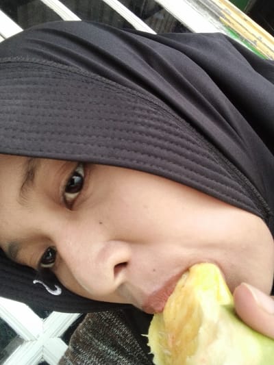 A close-up, low-angle shot captures a person wearing a black hijab and a brown knitted sweater, with their face partially obscured as they eat a piece of bright yellow fruit. The person's right eye, a dark brown iris with a reflective highlight, is visible. Their lips are slightly open, engaged with the fruit. A white, curved mark, possibly a logo or stitching, is present on the black fabric of the hijab near the person's cheek. The background includes a white criss-cross patterned window frame and some blurry green and yellow elements. The image appears to have been taken outdoors, possibly on a porch or near a window, in the city of Benuaraja, Indonesia.