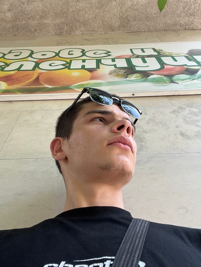 A low-angle shot in Karlovo, Bulgaria, captures a young male looking upwards. He has short, dark hair, minimal facial hair, and sunglasses perched on his head, reflecting the sky. He wears a black t-shirt with a white partial logo visible. A dark grey, textured shoulder strap crosses his right shoulder. Behind and above him, a horizontal sign features white and green Cyrillic text, including "добе" and "счу", overlaying images of sliced oranges and various green vegetables such as lettuce, broccoli, and pickles. Above the sign and to the sides, a textured, light tan wall is visible. A single green leaf hangs into the frame from the upper right corner.