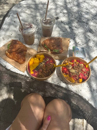The image provides a top-down view of an outdoor meal, likely breakfast or brunch, set on a light-colored, textured stone or concrete surface. The scene is bathed in sunlight, evidenced by prominent dappled shadows cast by overhead foliage.
The meal consists of several items arranged across the surface. Two large, light brown paper or cardboard bowls are filled with colorful fruit and granola. The upper bowl contains a mix of diced fruits including watermelon, mango, strawberries, banana, and possibly kiwi, generously topped with granola and various seeds (such as pumpkin seeds or chia seeds). A bright yellow spoon is stuck into this bowl. The lower bowl features a vibrant pink smoothie base (suggesting dragon fruit or a berry blend), topped with neatly cut mango pieces, granola, and chia seeds. This bowl also contains a yellow spoon.
To the left of the food bowls, there are two clear plastic cups holding iced beverages, likely iced coffee or a similar milky drink. Both drinks are topped with a layer of cream or foam and sprinkled with a dark powder, possibly cocoa or cinnamon. Each cup has a light brown, eco-friendly straw (appearing to be made of paper or bamboo) inserted.
Below the upper iced drink, a golden-brown, flaky croissant is placed on a piece of brown paper. Green lettuce is visible within the croissant, suggesting it's a sandwich with a savory filling. Another identical croissant, also on brown paper, is situated below the lower iced drink.
Near the upper bowl and above the upper croissant, a small, white rectangular pouch or sachet is visible. It has green and blue graphic elements and the brand name "Magimix" is partially legible on its front, accompanied by what appear to be recycling or eco-friendly symbols.
On the far right side of the image, part of a person's lower body is visible, specifically their thighs and knees, suggesting they are seated next to the table. Their skin appears tanned. A hand with brightly painted pink fingernails rests on one of the knees. A dark, rough-textured surface, possibly a wooden bench or a tree trunk, runs vertically along the right edge of the frame, separating the food from the person's legs.
The overall impression is that of a healthy and appealing outdoor meal enjoyed on a sunny day.