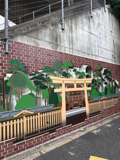 This image presents an outdoor urban scene, likely viewed from an elevated perspective, showcasing a blend of industrial architecture, public art, and street infrastructure.
On the left, a large, textured concrete wall or support structure is visible. It's grey, marked with rectangular divisions and signs of weathering, and features several metal bolts and fixtures. A robust metal scaffolding or fence structure is attached to the far left side of this concrete surface.
Adjacent to the concrete, a prominent vertical surface decorated with a deep red brick pattern dominates the central area. Adorning this brickwork is a large, stylized mural depicting a dense forest scene. The mural features numerous trees and foliage in various shades of green, outlined in gold, against a lighter, soft grey-blue background, creating a layered and somewhat whimsical aesthetic.
Integrated with or in front of this mural is a light-colored wooden structure, resembling a traditional Japanese torii gate, with its distinctive crossbeams and uprights. Below the torii gate, part of the mural depicts a building facade with windows and doors, suggesting a shop or house. Further right along the brick-patterned wall, another wooden structure, composed of horizontal slats, extends, possibly acting as a decorative fence or seating.
The far right of the image reveals a stretch of dark asphalt road, bordered by a concrete curb and a yellow tactile paving strip, commonly used for the visually impaired in Japan. The overall lighting suggests daytime, likely overcast, as there are no harsh shadows. No people or visible text are present, and the scene depicts a static urban landscape featuring decorative public art. The architectural and street elements are consistent with an urban setting in Shibuya, Japan.