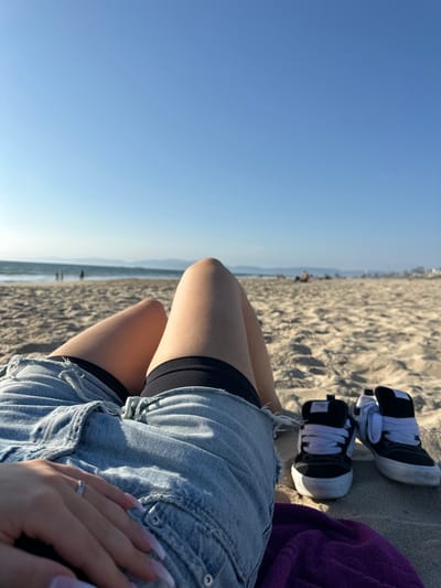 An individual is depicted reclining on a light brown sandy beach in Manhattan Beach, California, United States, under a clear, bright blue sky. The viewpoint is from the person's perspective, looking towards the Pacific Ocean. The lower torso, legs, and left hand of the individual are visible. They are wearing light blue denim cutoff shorts over black compression shorts. A silver ring is present on the ring finger of the left hand, which features French-tipped nails. A pair of black and white athletic shoes with white laces are placed on the sand to the immediate right of the individual's legs, alongside a partially visible purple towel. In the midground, the ocean shows small waves, and several indistinct figures are visible walking or resting on the beach and near the water's edge. A hazy landmass is discernible on the distant horizon, with faint coastal structures visible further to the right.
