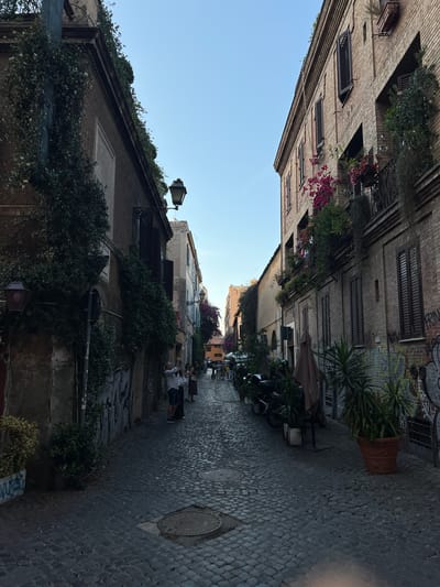 The image depicts a narrow, historic cobblestone street or alleyway, characteristic of an old European city like Rome, under a clear blue sky during the daytime. The street is lined on both sides by multi-story buildings, featuring facades of brick and stucco with numerous windows often fitted with dark wooden or metal shutters.
Abundant greenery decorates the scene, with ivy climbing walls, lush plants in window boxes and on balconies, and vibrant pink flowering plants (resembling bougainvillea) adding splashes of color. Large potted plants, including a prominent yucca-like plant in a terracotta pot, are placed along the street. Traditional lampposts are visible, providing ambient lighting.
The cobblestone pavement is well-maintained and features several circular manhole covers. On the right side of the street, a few tables and chairs are set up under a partially open brown umbrella, indicating an outdoor dining area or cafe. Several motor scooters or mopeds are parked alongside the building on the right.
A few people can be seen further down the street on the right, some seated and others standing casually, suggesting everyday activity. The overall atmosphere appears calm and pleasant. Graffiti is visible on the upper section of the right-hand building, with parts of the word "GRAF" discernible among other markings.