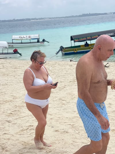 The scene depicts a beach in Zanzibar, Tanzania, under an overcast sky. In the foreground, a middle-aged woman with short, light-colored hair and sunglasses stands barefoot on light-colored sand, angled slightly right. She wears a white two-piece swimsuit and is engaged with a black smartphone held in both hands, looking down at its screen. Sand adheres to her feet; her left big toenail is painted pink. To her right and slightly behind, a bald, shirtless middle-aged man walks towards the right side of the frame. He wears light blue swim trunks with a white geometric pattern and carries a clear plastic cup filled with light-colored snacks in his right hand. He looks downwards.
The background features several small, motorized boats moored in shallow, clear turquoise water, which transitions to a deeper blue further from shore. One boat displays "GLTR" on its white canopy, another bears "77751904" and "321" on its hull, and a third, painted green, yellow, and blue, has "NO PAIN NO GAIN" written on its dark canopy, with two orange life preservers attached to its side. A faint landmass with visible structures is present on the distant horizon. Minimal natural debris is scattered in the water and on the immediate shoreline.