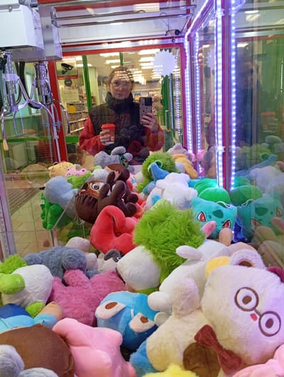 A claw machine filled with plush toys, including characters resembling Bulbasaur and other various creatures, is the primary focus of the image. The machine's interior is illuminated with LED strips. The reflection in the machine's glass shows a person holding a smartphone and a small orange container, seemingly taking a picture. The person has short, curly hair, is wearing glasses, and is wearing a black jacket over a red shirt. Behind the person's reflection are posters and merchandise displays. The location is Yurgamysh, Russia.