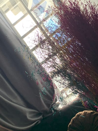 The image shows a close-up of a decorative hanging arrangement with thin, colorful strands (predominantly shades of purple, green, and gold). The strands are dense at the top and more scattered towards the bottom. In the background, a window with a grid-like pattern is visible, with bright light shining through. The arrangement appears to be positioned indoors, casting shadows on a smooth, dark gray or black material that drapes downwards to the right of the frame, this could be draped over the back of a seat cushion, a part of which is visible to the upper right. It is impossible to verify that this image was taken in Ibasa, Nigeria.