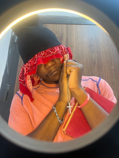 An individual wearing a black beanie and red bandana is lying on a pink garment, with a red book resting on their arm. A silver cross necklace is held in their hands. A pink wristband is visible. The location is undetermined.