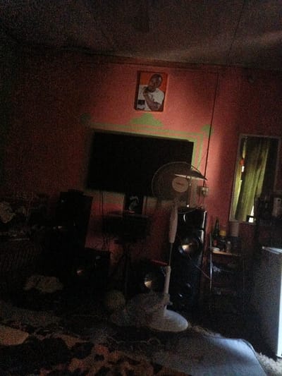 The image depicts a dimly lit indoor residential scene, presumably within the city of Gwagwarwa, Nigeria. The room features reddish-orange painted walls, with visible green paint splashes or markings above a television screen.
Centered on the upper portion of the right wall is a framed photograph of a light-skinned man wearing a white t-shirt, looking towards the left and holding his hand to his chest, appearing to smile. Directly beneath this photograph, mounted on the wall, is a dark, rectangular flat-screen television. To the right of the television, a white Binatone stand fan with visible dust on its blades is positioned, partially obstructing a large, dark speaker system located on the floor.
Further to the right, a rectangular mirror is affixed to the wall, reflecting a window with light green curtains. Below the mirror, a dark shelf unit holds various indistinct objects. On the far right, a tall, light-colored appliance, possibly a refrigerator or cooler, is visible.
The floor is covered with a patterned rug or carpet. The overall lighting is low, contributing to a grainy appearance in the photograph, with shadows obscuring significant detail in the lower left and central foreground. No individuals are present, and no actions or interactions are occurring within the frame. The environment suggests a lived-in space with personal effects and entertainment equipment.