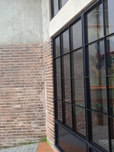 The image shows a corner of a building with a large window. The window is made of black metal framing and has multiple panes of glass. The building is constructed of brick with a concrete finish on the top portion. The brickwork is brown and weathered, while the concrete is light gray. The window reflects the exterior of the building, including a portion of the brick wall. The reflection in the glass shows a small potted plant on the left side of the window and a larger plant on the right side. The location of the building, which can be determined from the coordinates provided, is in the city of Surakarta, Indonesia.